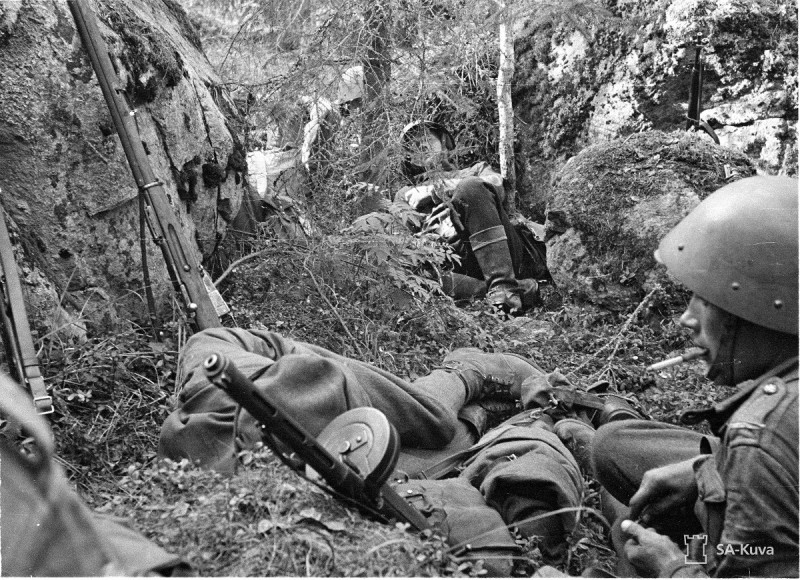 Finnish soldiers resting before an attack. An older Mosin-Nagant rifle (indicated by its curved rear sight leaf) is leaned against the earth on the left. Date taken: July 30, 1941.